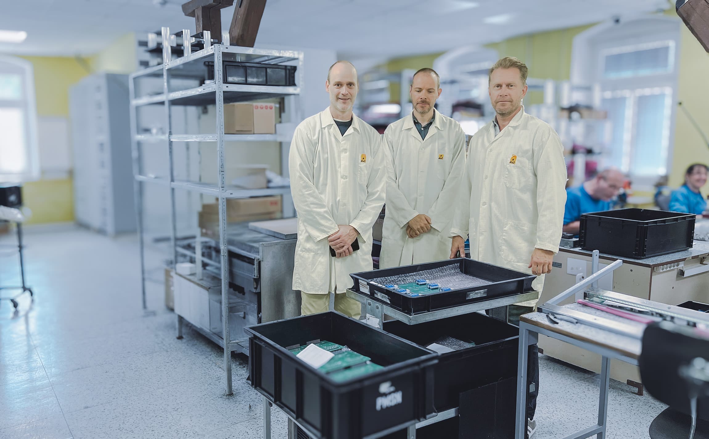 Three engineers in ESD-safe lab coats in an electronics production and assembly room.