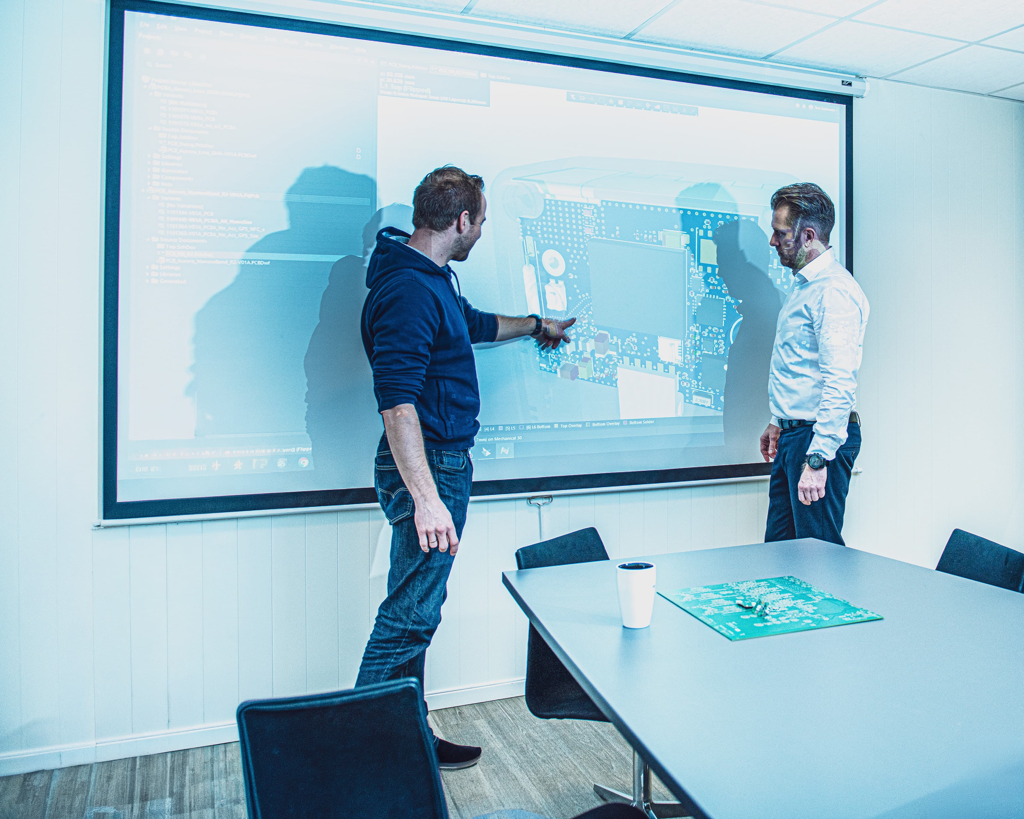 Two engineers reviewing a PCB design on a projector screen in a meeting room.